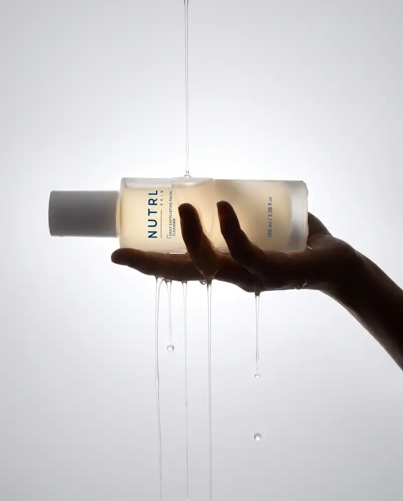 Hand holding a NUTRL Daily Exfoliating Facial Cleanser bottle under a stream of water